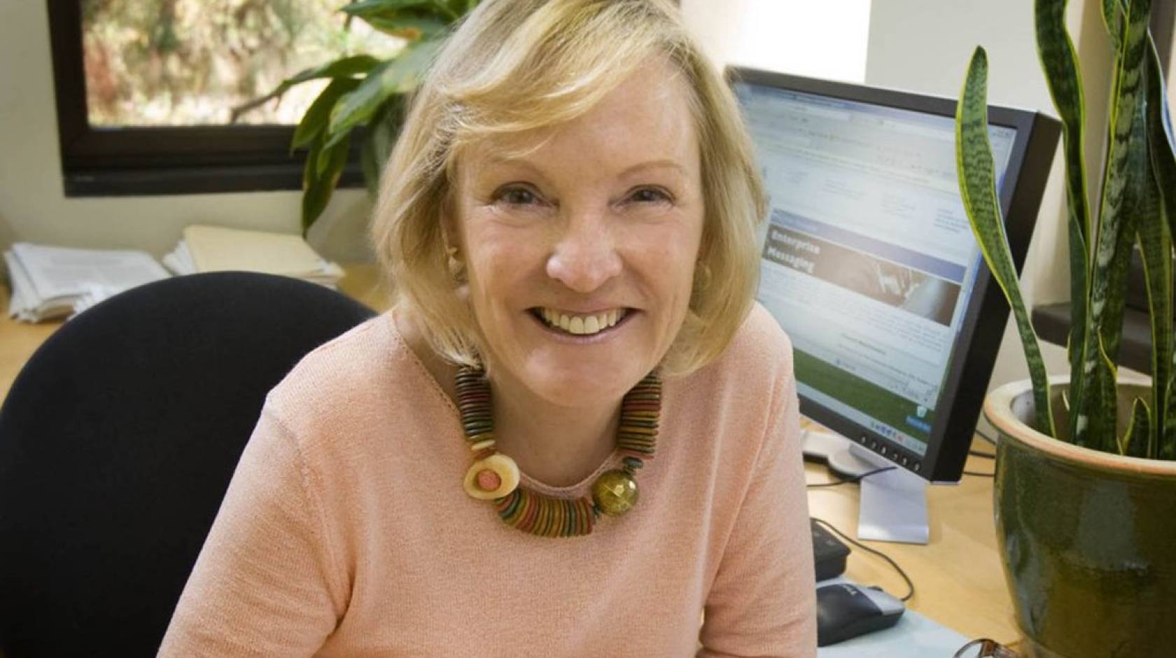 Shelley Taylor, white woman with blonde hair cut in a bob, wearing a pink sweater, smiles at the camera sitting at her desk, with a monitor and a potted planted in the background.