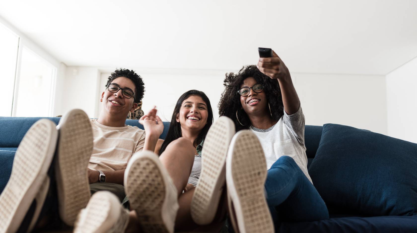 Three smiling teenagers sitting close together on a couch, one is holding a TV remote