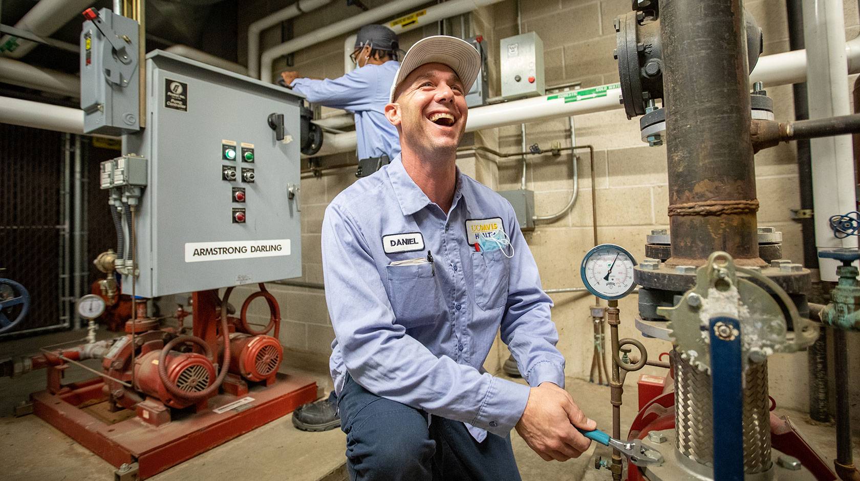 A maintenance worker in a light blue uniform kneels in a mechanical room, smiling as he uses a wrench to adjust a large pipe valve, while another worker operates a control panel in the background surrounded by pumps, gauges, and industrial piping.