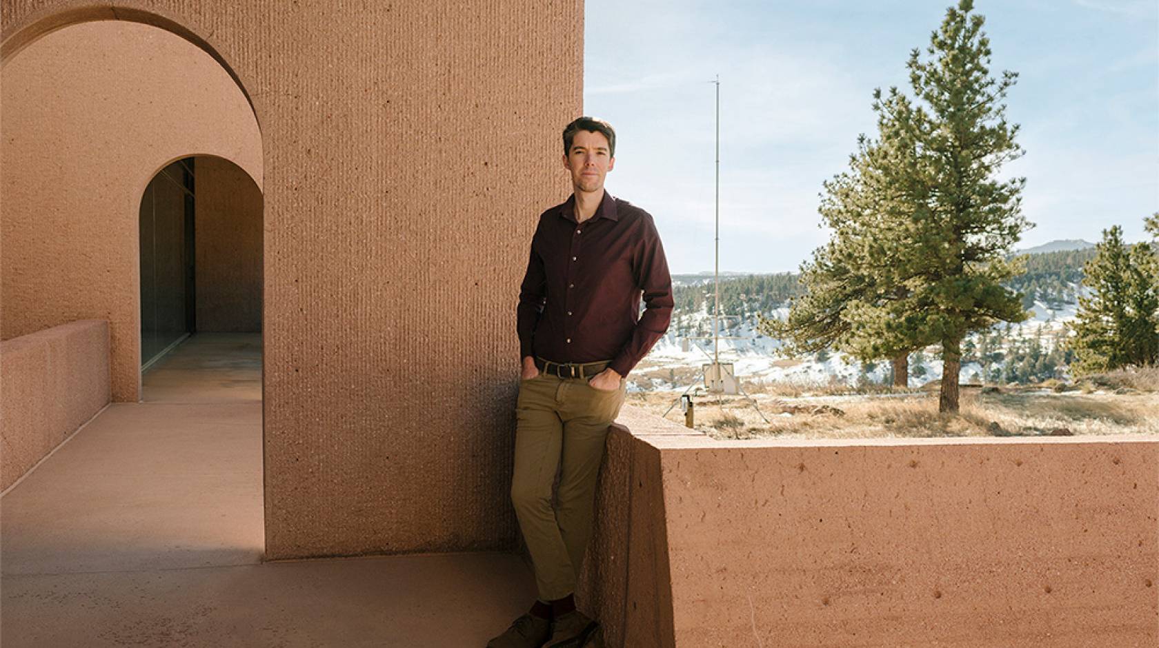 Daniel Swain, wearing khakis and a maroon shirt, leans against a low adobe-colored wall, with a snowy forest in the backdrop and an arched breezeway behind him. 