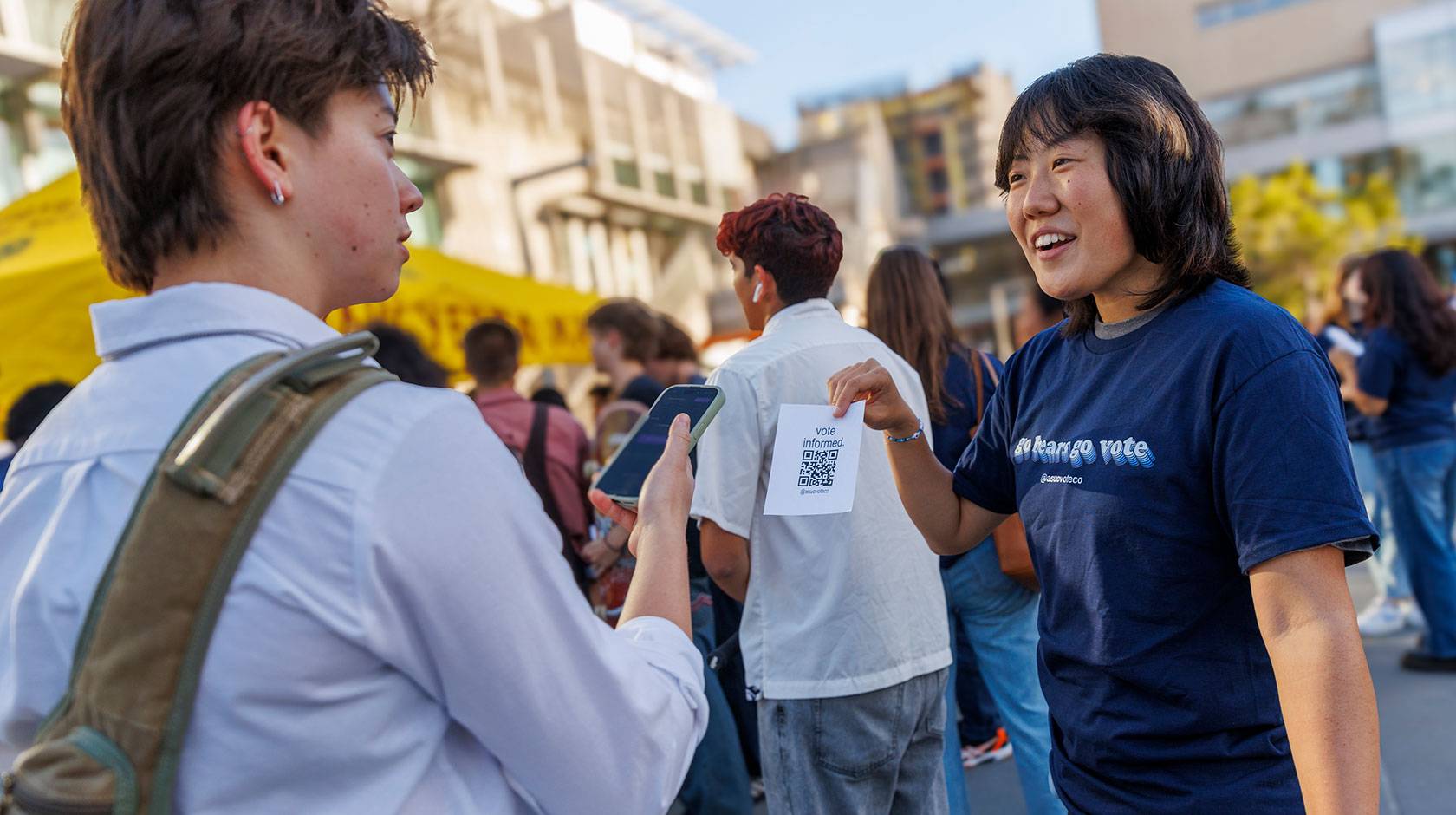 Student wearing a blue shirt reading GO BEARS. GO VOTE. holds a paper with a QR code printed up for a fellow student to scan, on campus at UC Berkeley