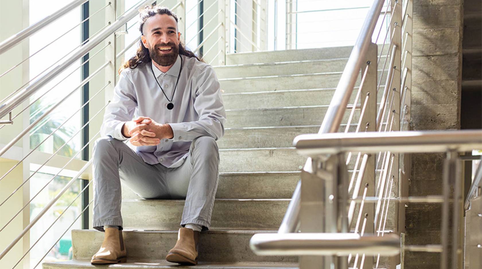 Trevor Auldridge-Reveles sits in a concrete and steel staircase, leaning elbows on legs, smiling