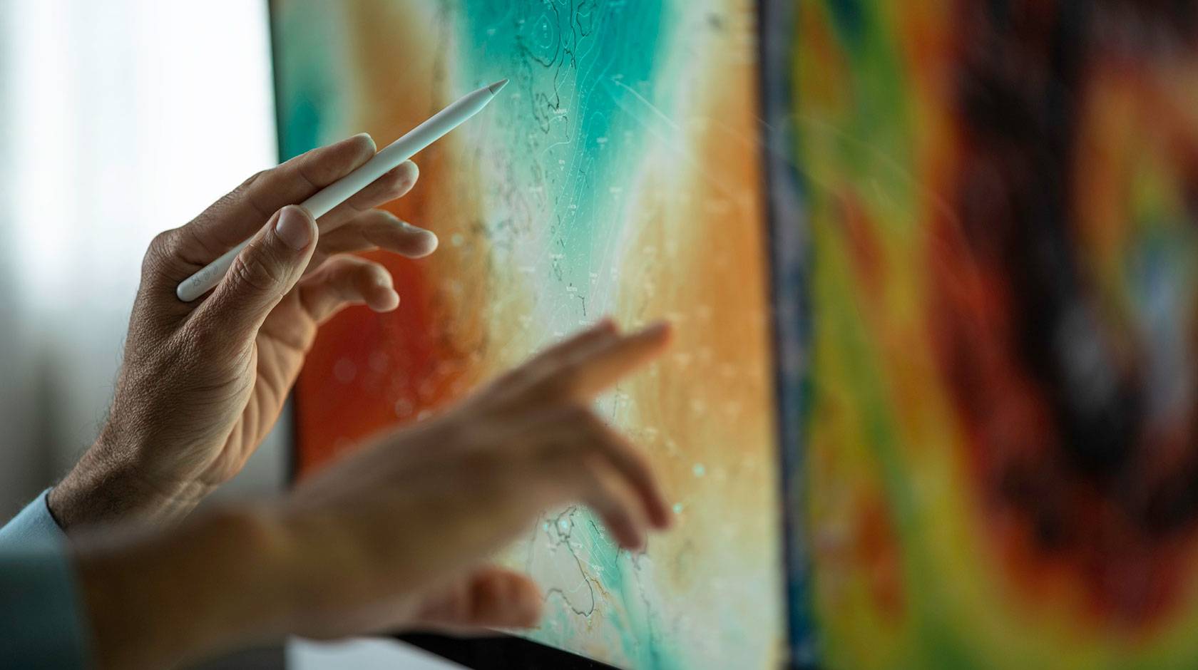 Hands using a stylus and finger to interact with a large touchscreen displaying a colorful weather map