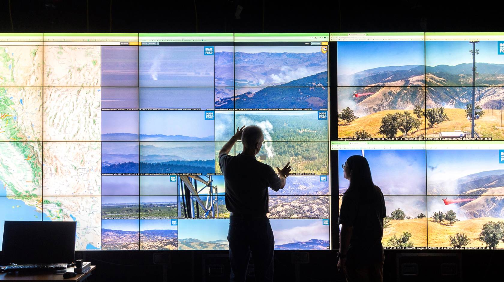 Two people in silhouette look at a large series of screens with visuals of different California landscapes, and a topographical video map of California to the left