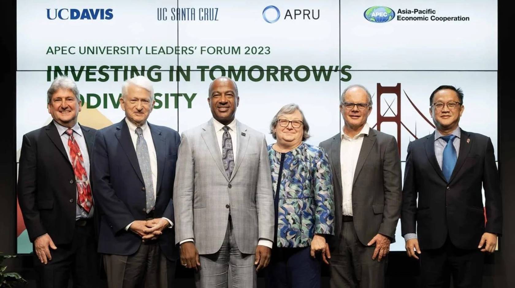 6 people in business attire stand shoulder-to-shoulder and smile for the camera in front of a video screen background reading "APEC University Leaders Forum: Investing in Tomorrow's Biodiversity"