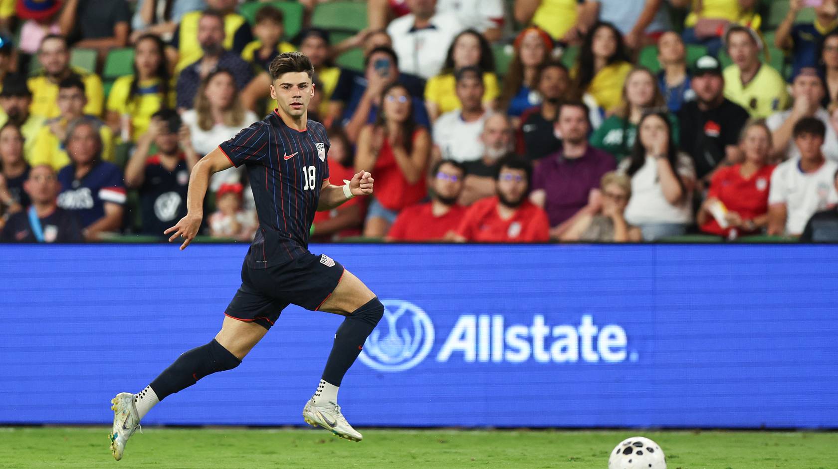 Max Arfsten of the United States in a navy blue uniform with stripes drives a ball during an international friendly match in front of the crowd, looking toward the camera