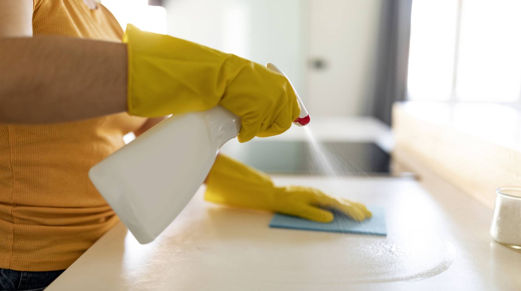A woman wearing a peach shirt and yellow rubber gloves uses an unmarked spray bottle to clean a kitchen counter with a rag.