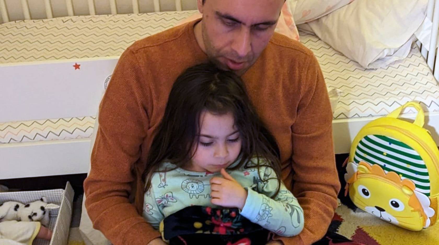 A balding man holds a young daughter as they read together, looking at a screen