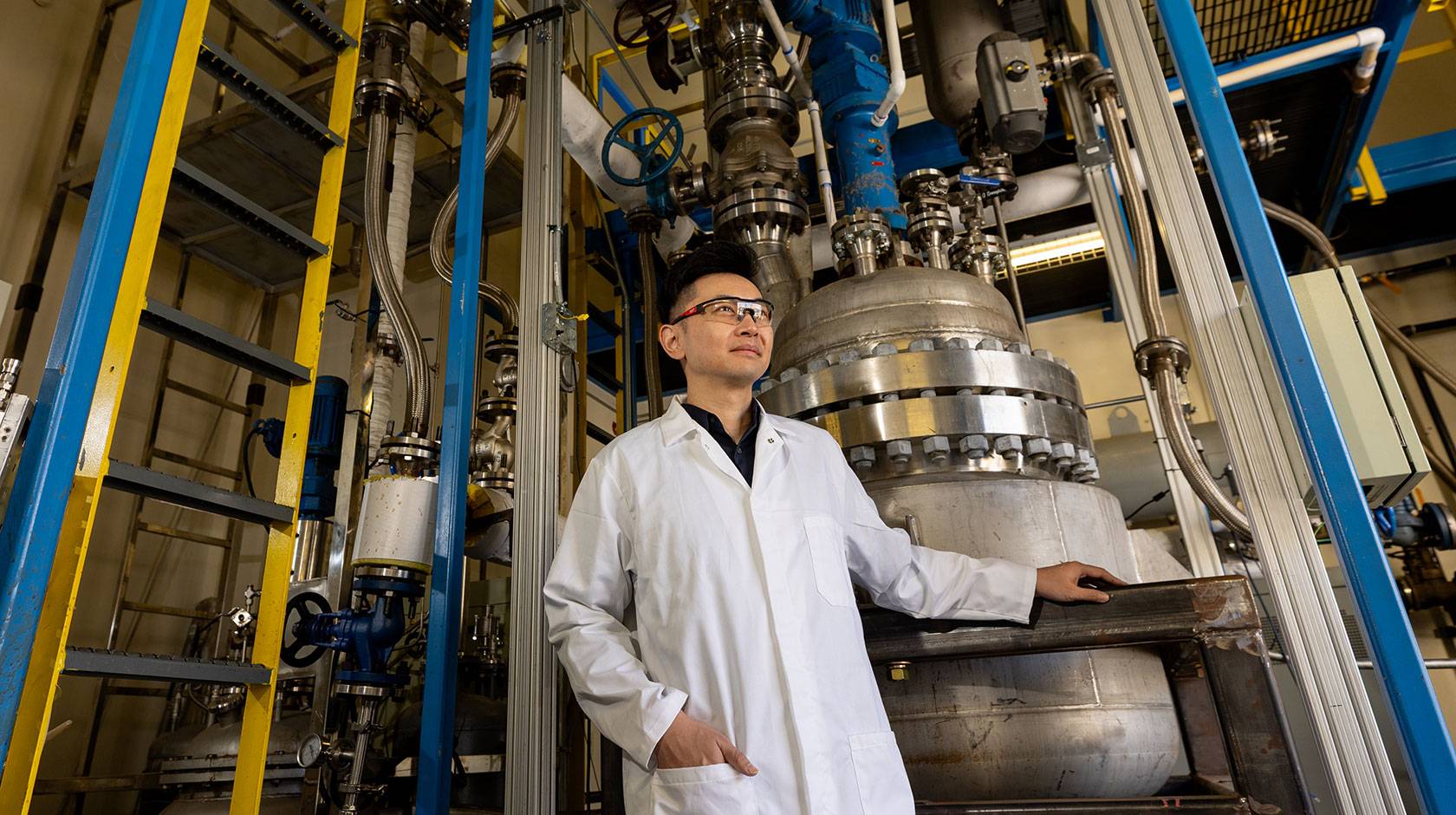 A man wearing a white lab coat and safety glasses stands in an industrial laboratory next to a large stainless steel machine with pipes and bolts, resting one hand on a metal railing.
