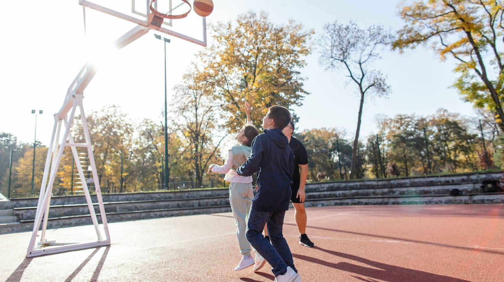 Two kids play outdoors on a basketball court with a parent