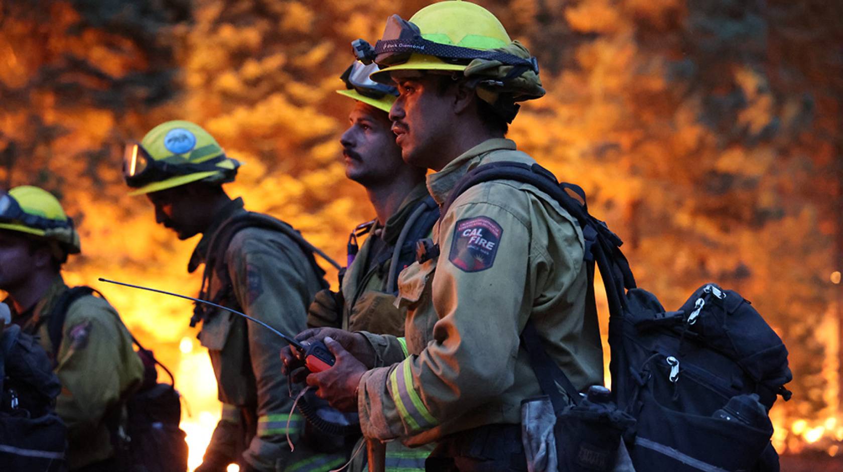 Four firefighters stand against a backdrop of flames. The scene is dark, and 2 young male firefighters hold a device with a long antenna