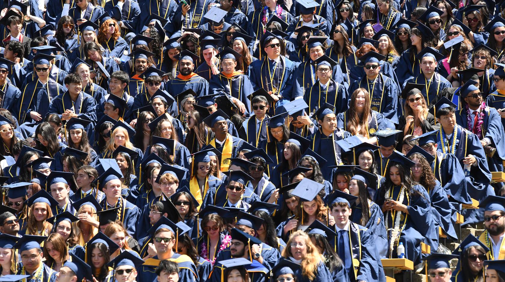 A high-angle shot of a dense crowd of university graduates in blue caps and gowns filling stadium bleachers, accented by colorful leis, stoles and sunglasses under bright sunlight.