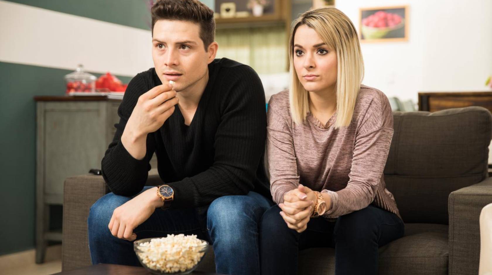 A white man and a white woman watching TV together, looking somber