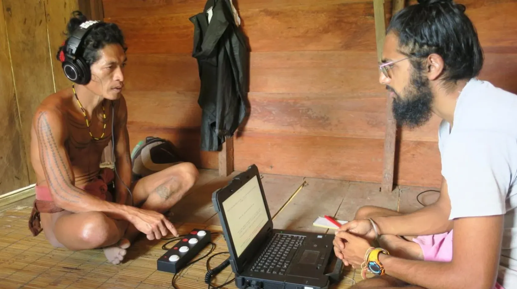 An Indigenous man sits on the floor listening to headphones, facing a researcher with a laptop