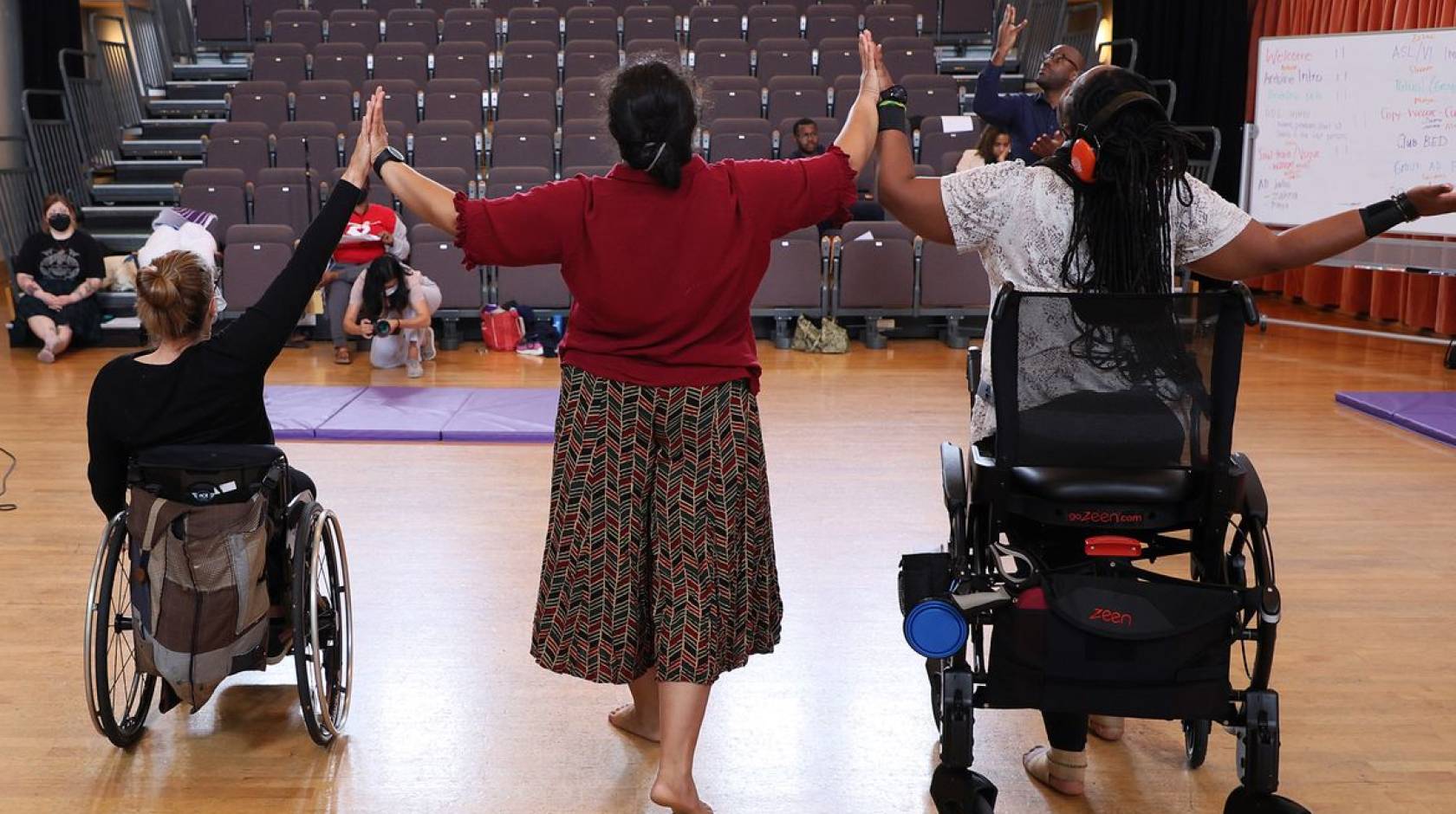 Three disabled dancers, photographed from behind, touch hands while raising their arms.