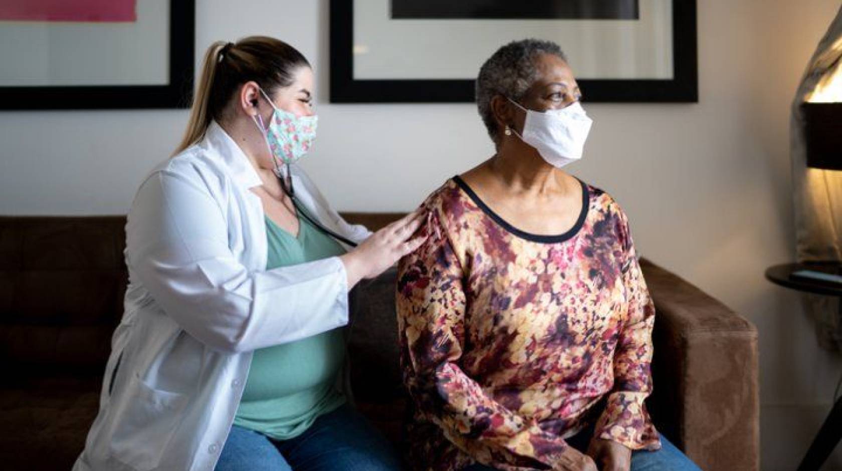 Doctor examining an older Black woman's breathing, both wearing masks