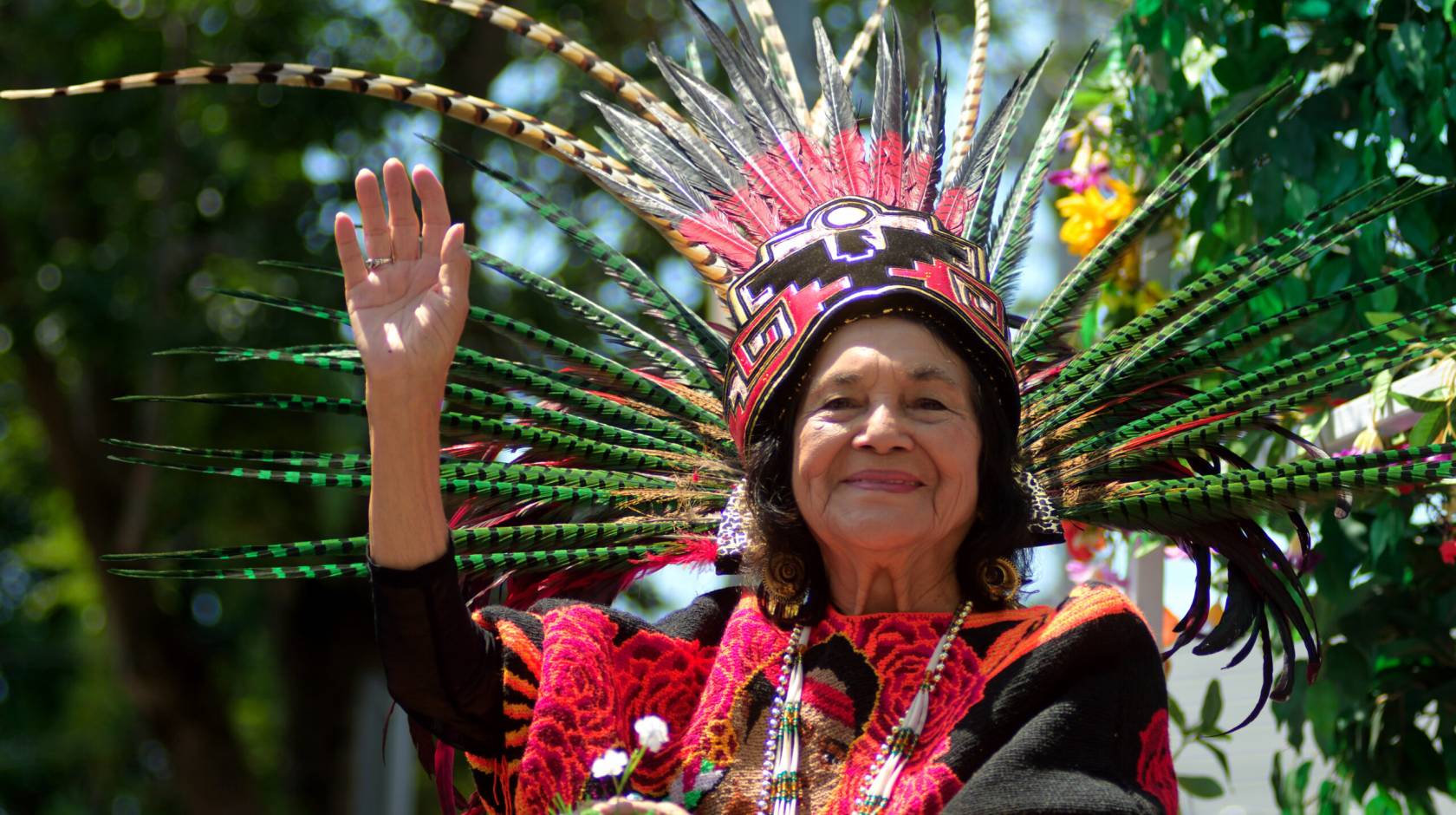 Dolores Huerta in traditional clothing and a headdress waves and smiles