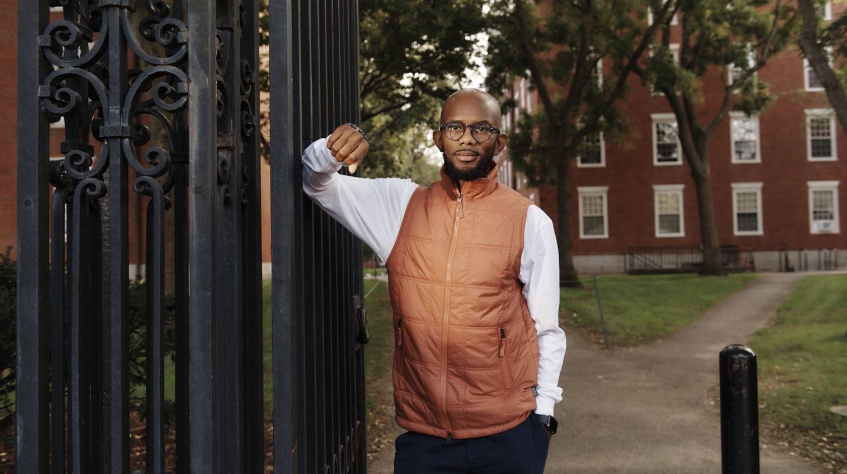 Young Black man in an orange vest and glasses standing against a gate on the Harvard campus