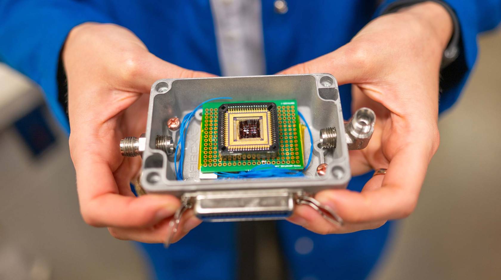 A pair of hands holds a small plastic box that contains a complex electronic chip