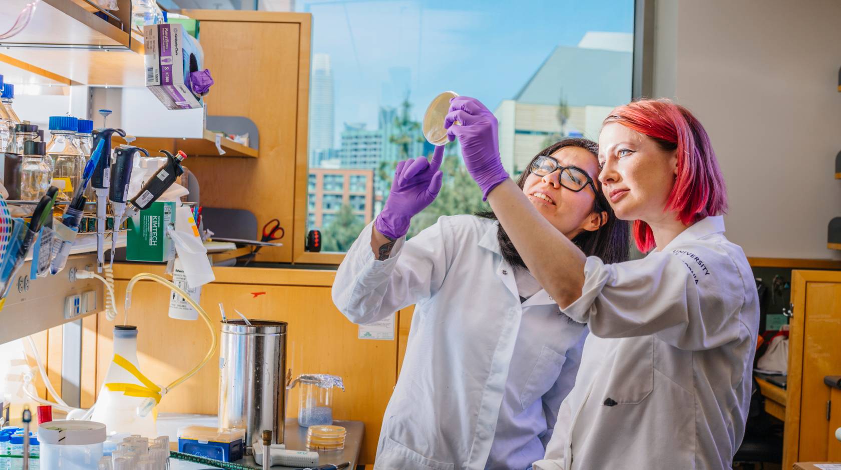 Two female students, one with pink hair, hold up a petri dish in a lab