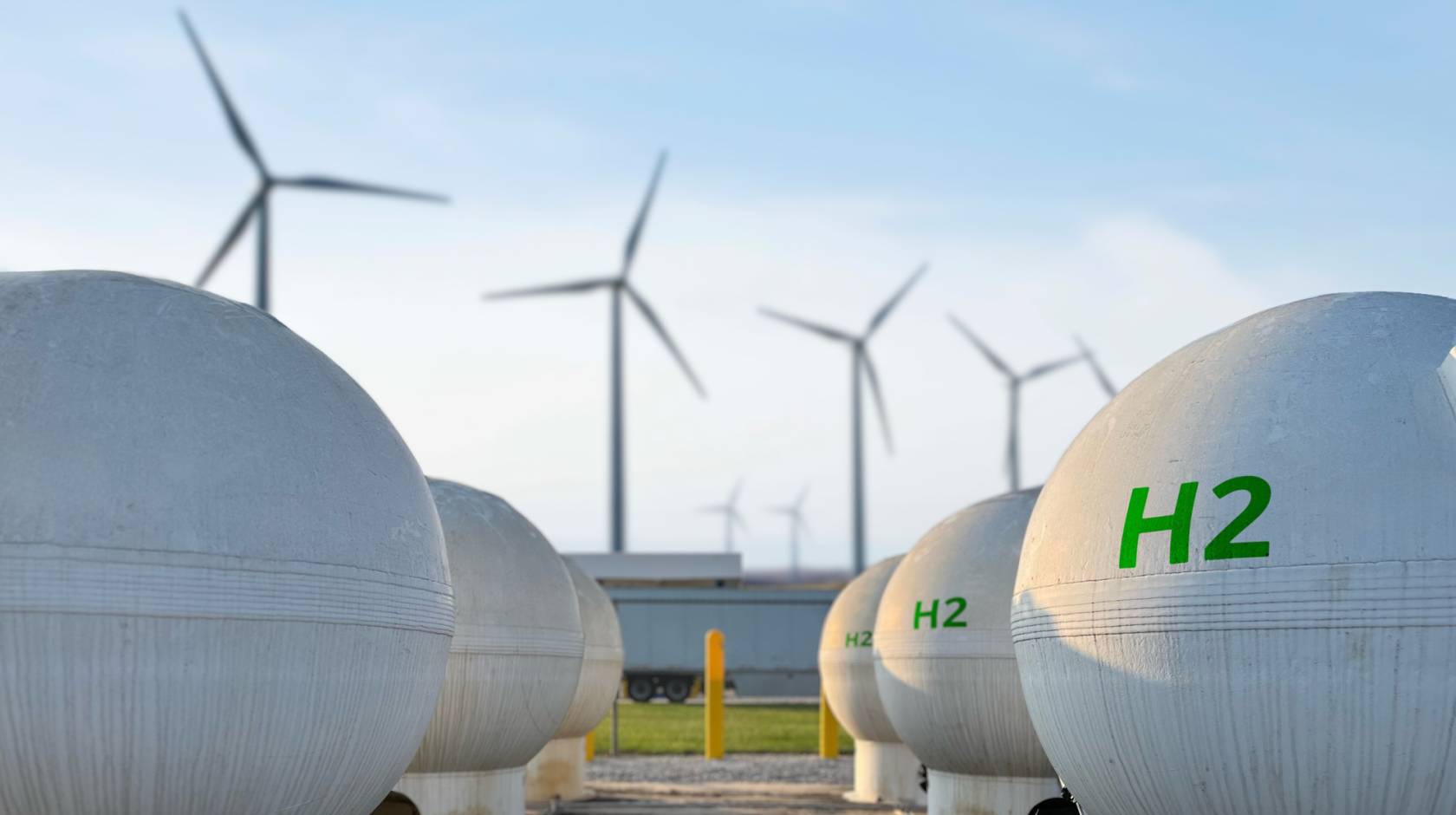 Two rows of spherical white tanks with H2 printed on them in green, with four wind turbines in the background, against a blue sky