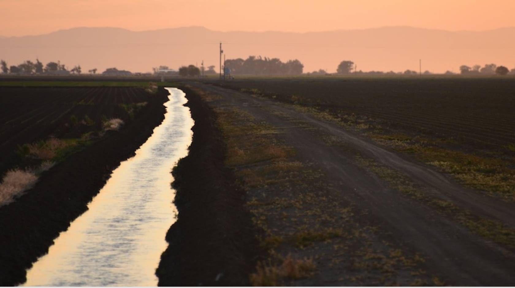 A irrigation canal runs away from the camera to a vanishing point. The fields on either side are dark, the canal reflects the orange-pink hazy sky