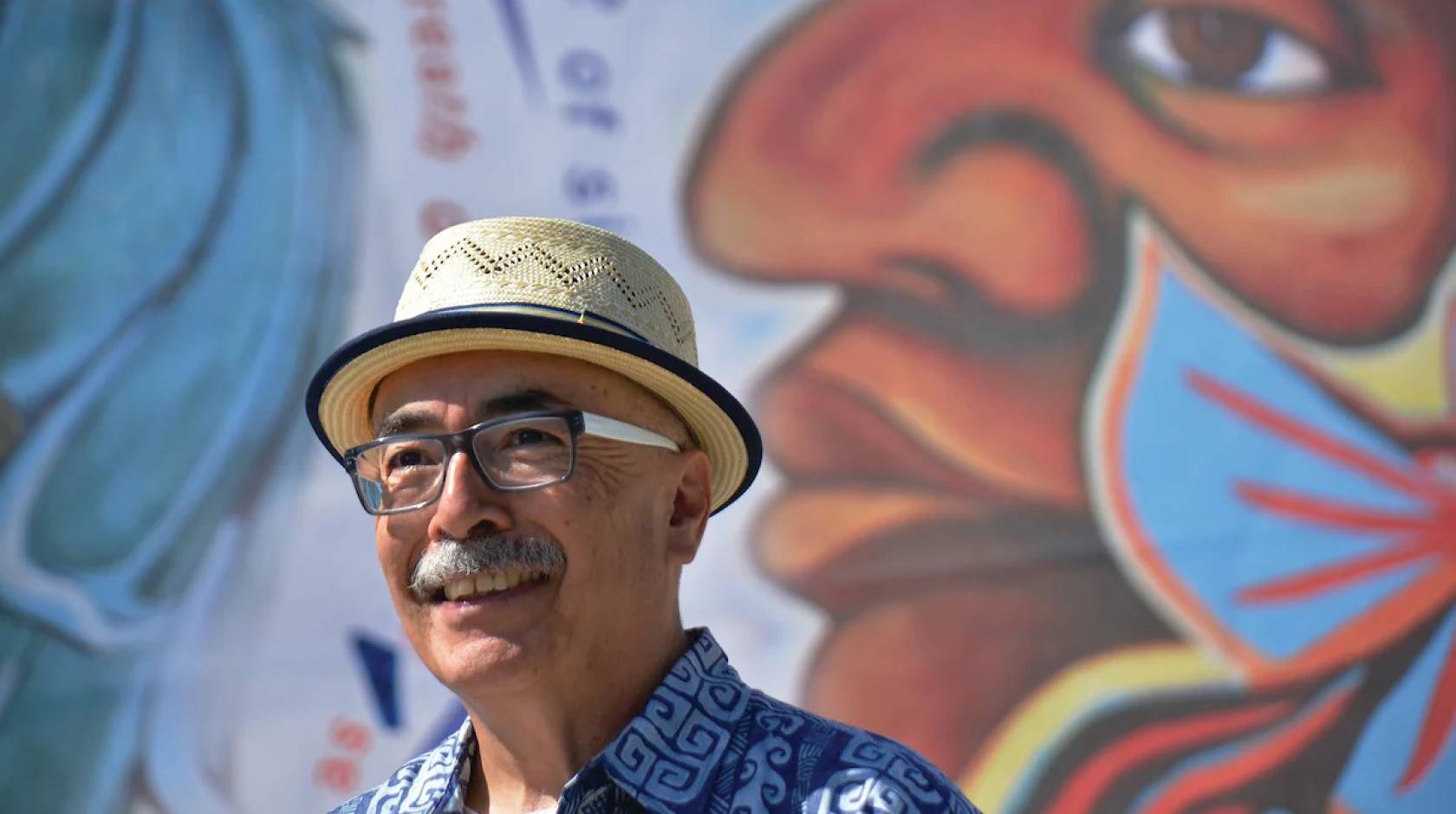 An older Latino man with a mustache, glasses and a jaunty hat in front of a colorful mural
