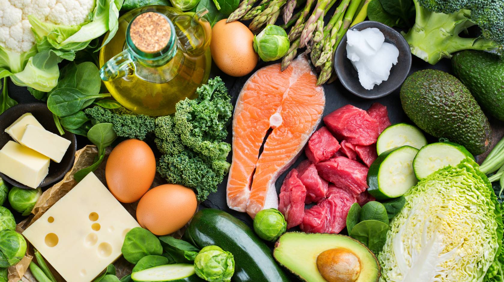 Assortment of healthy ketogenic low carb food ingredients for cooking on a kitchen table. Green vegetables, meat, salmon, cheese, eggs.