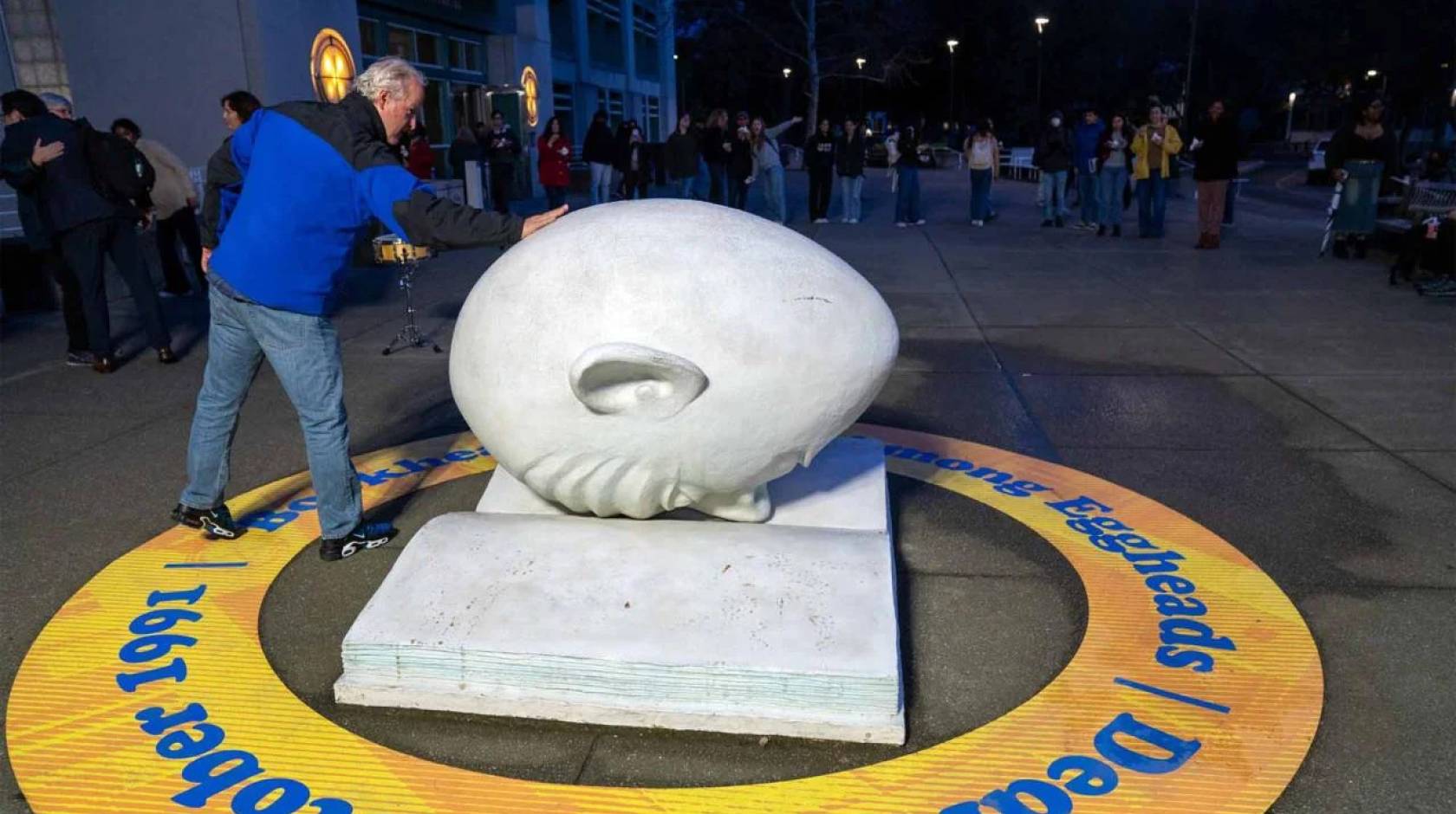 In the front of the photo, a man touches an egghead (a humorous but blank-looking human face shaped like an egg) which is faced down on a concrete open book; in the back of the photo, a line of people outside a museum at night