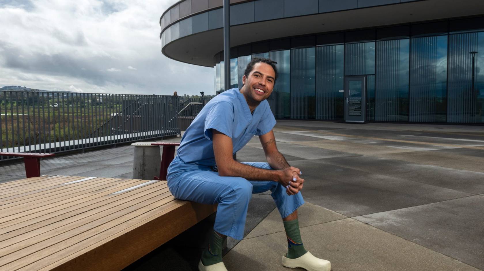 A person in blue scrubs sits outside a medical building and smiles