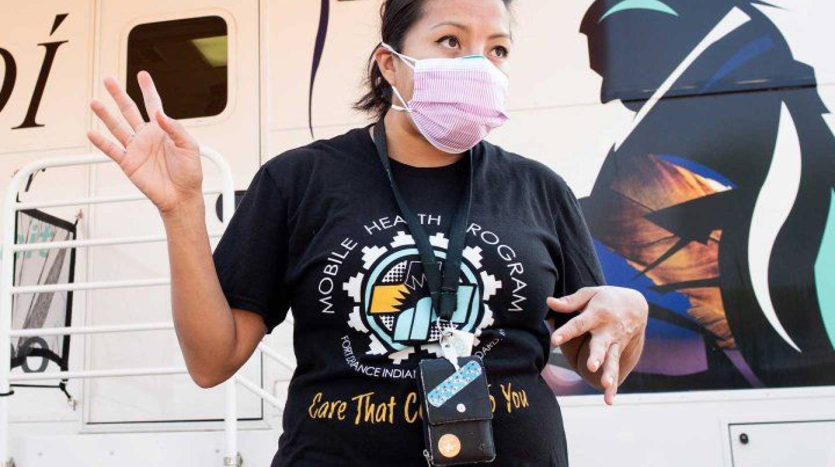 A young woman wearing a black t-shirt and a pink face mask gesticulates with her hands standing in front of a trailer