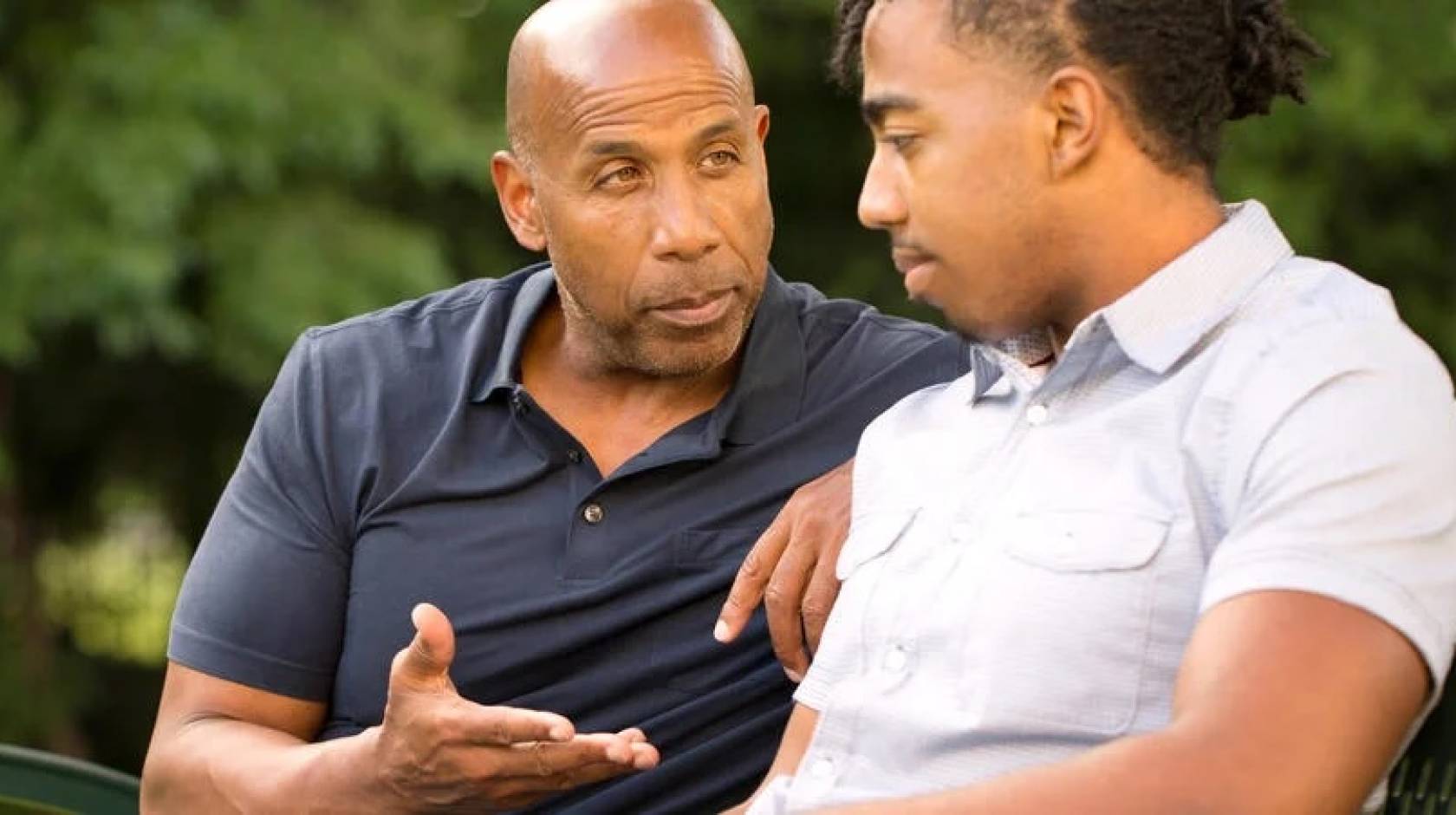 A father and his teen son sit on a park bench. The father is speaking intently to the son, who appears to be listening.