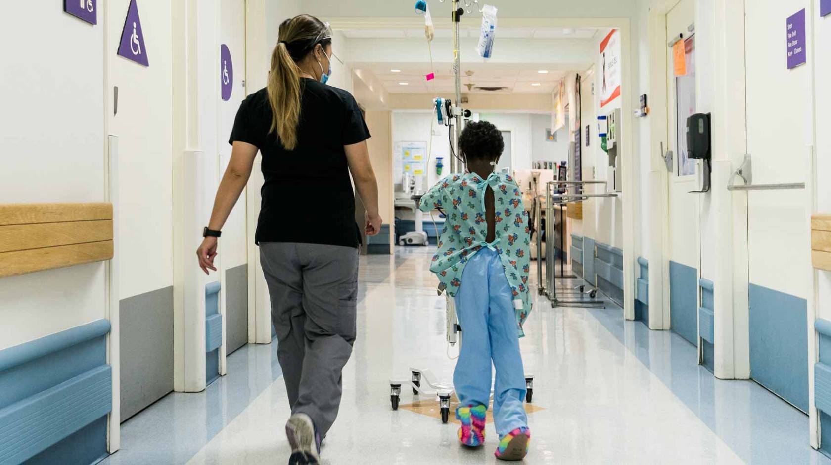 a nurse wearing a mask walks with a young Black patient around a hospital unit