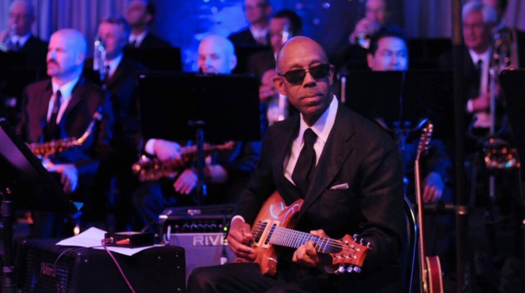 President Drake plays guitar on stage, wearing a black suit and sunglasses