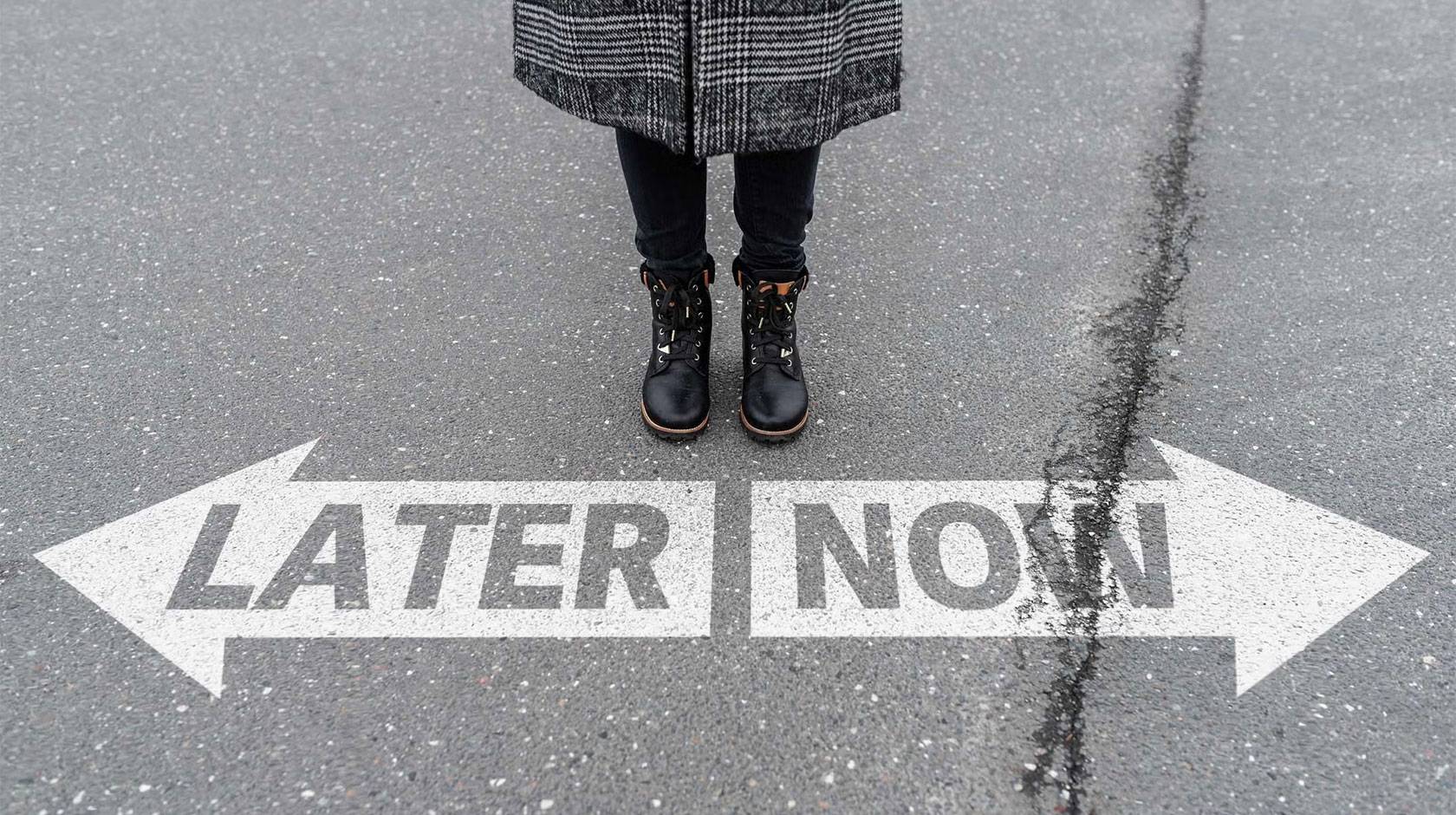 View of a person standing on pavement with only their legs and boots visible, positioned above two large white arrows painted on the ground—one pointing left labeled “LATER” and one pointing right labeled “NOW.”