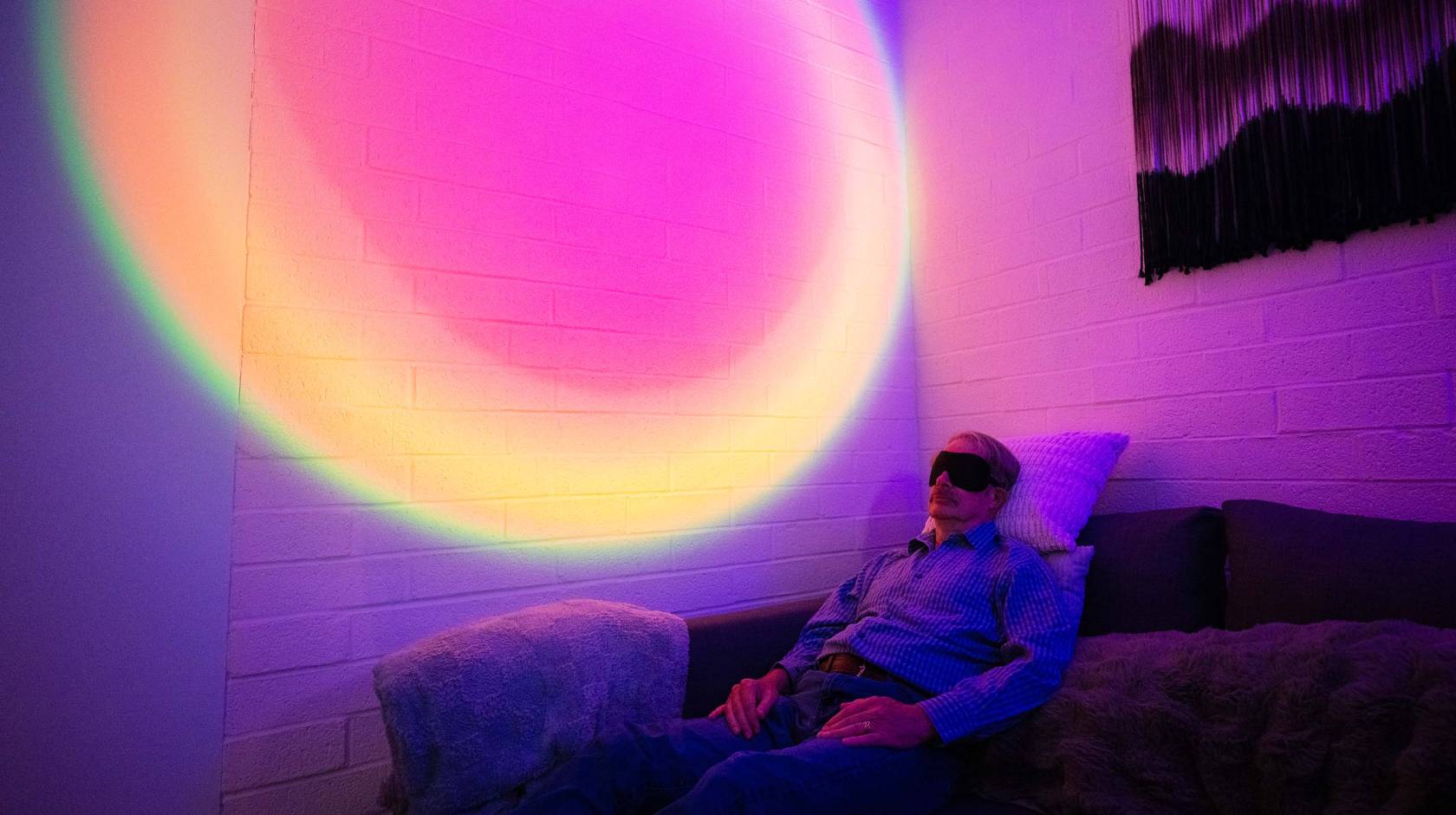 A man wearing an eye mask reclines on a couch in a darkened room. Behind him, the wall is illuminated with a circular, rainbow-colored light