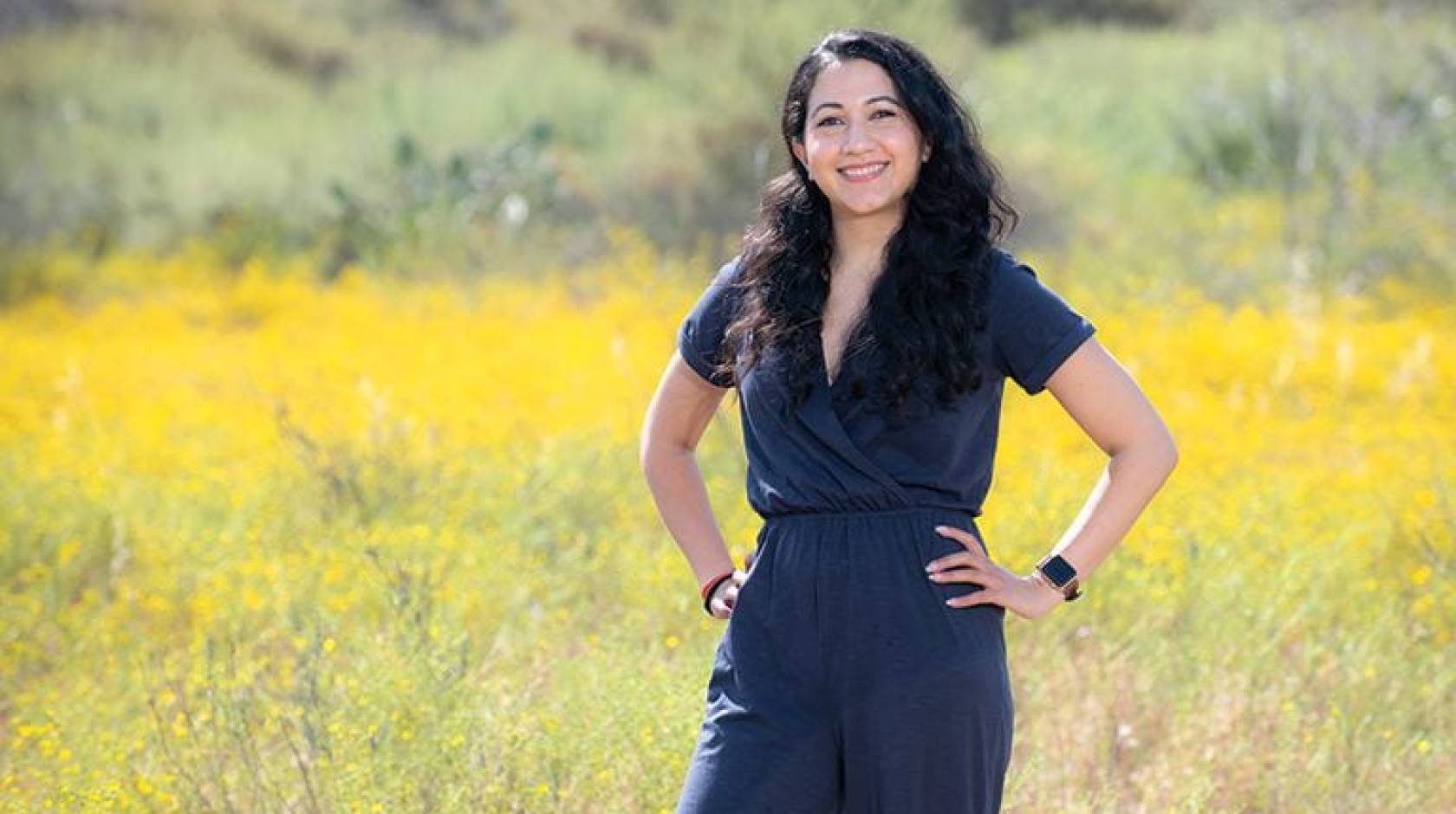 Woman with long dark hair, Shyla Raghav, standing in front of a yellow field of wildflowers