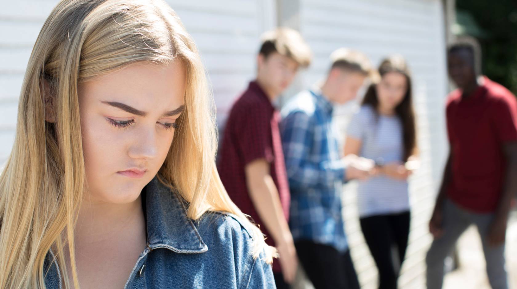 Blonde teenage girl frowns as peers behind her gossip