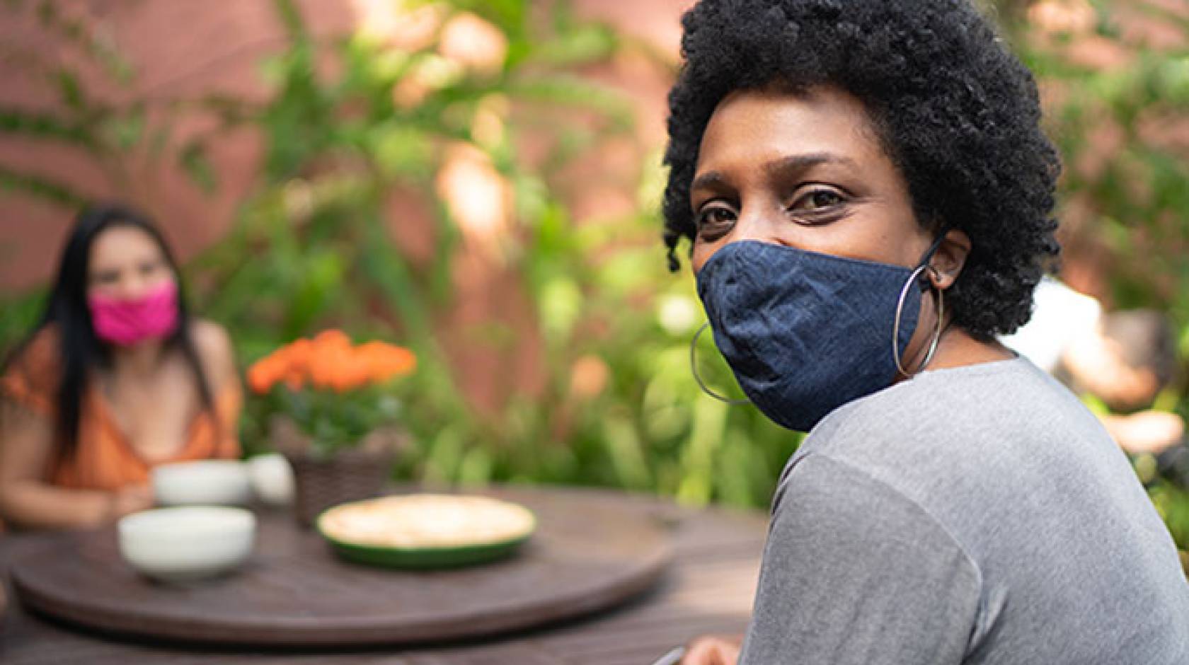 Black woman in mask looks at camera while socializing with friend outside