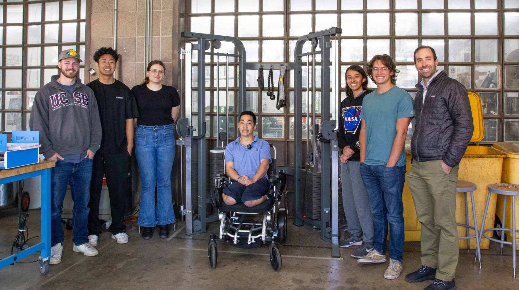 A team of students and a professor stands around a young man in a wheelchair underneath a workout machine