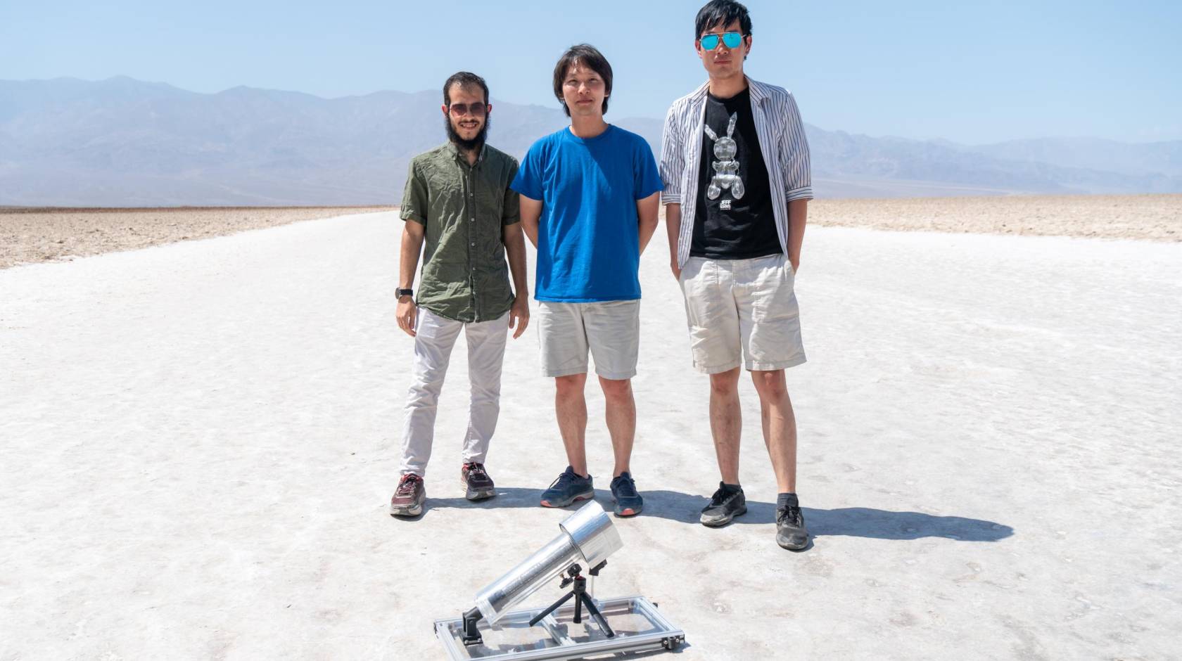 Three male students stand in white desert sands, behind a water harvester that looks like a microphone