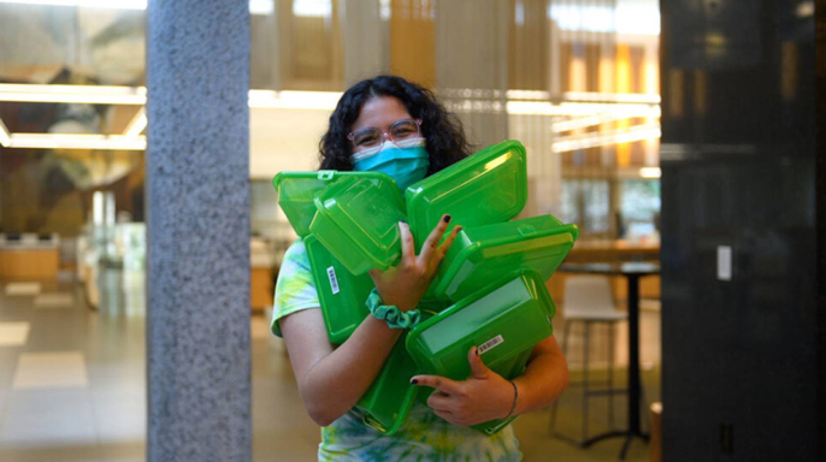 Woman in a mask carrying an armload of green reuseable food containers