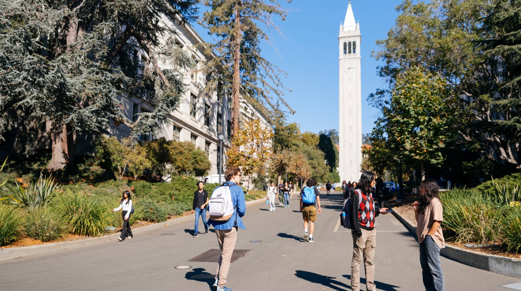 Students walk and talk on a tree-lined wide walkway in front of Sather Tower on the UC Berkeley campus