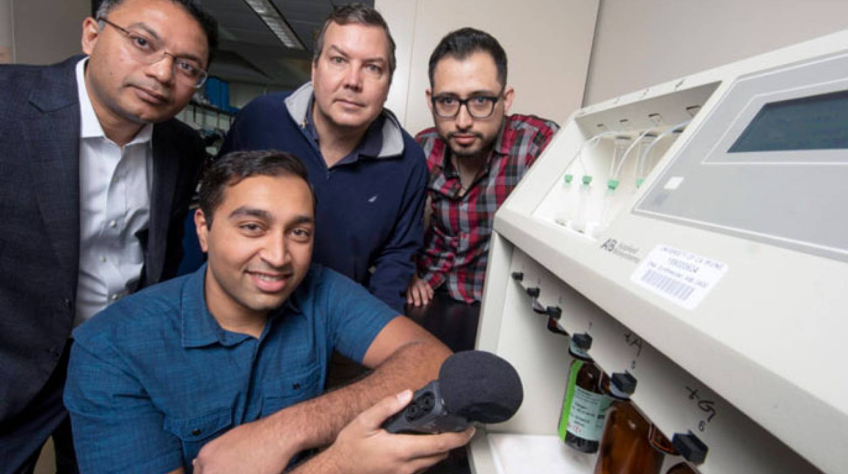 UC Irvine and UC Riverside researchers hold up a microphone to a DNA synthesizer