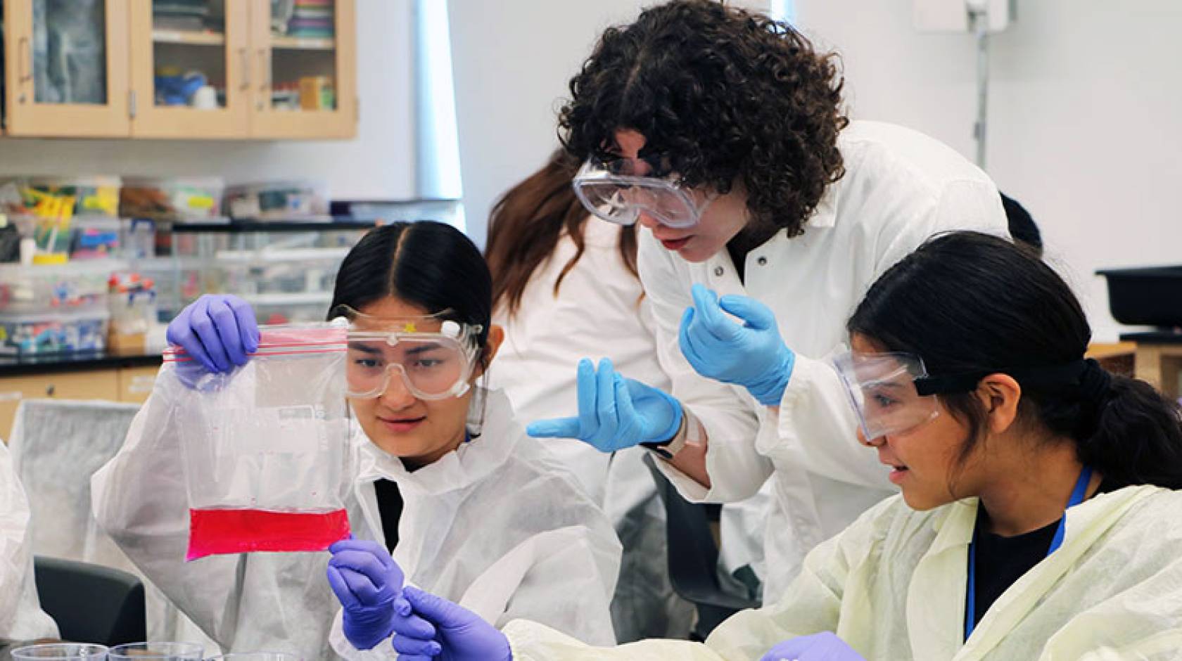 A college student in PPE shows two high schoolers in PPE how to collect a sample in a lab