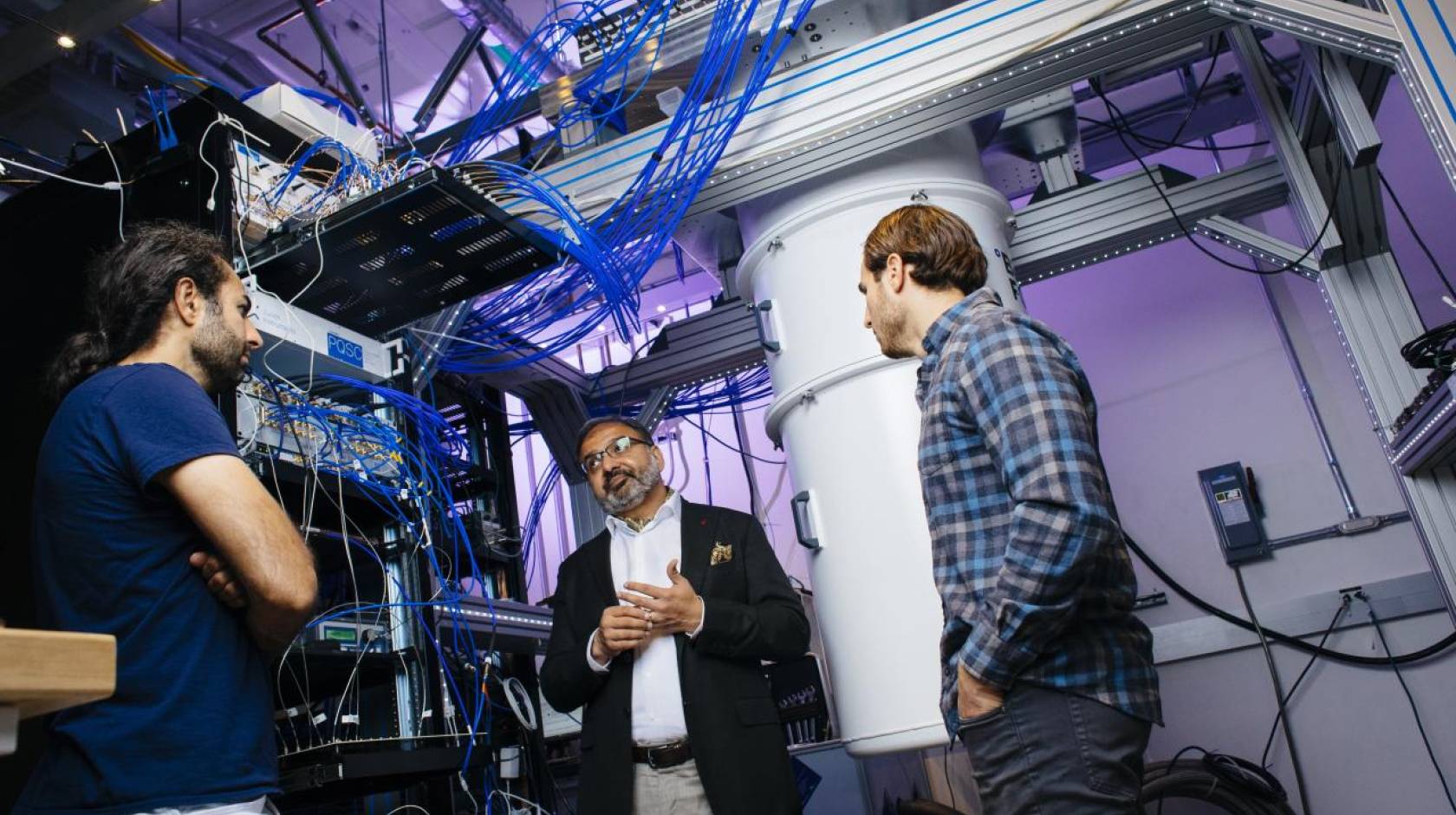 Three men talking in a quantum computing lab with purple lighting