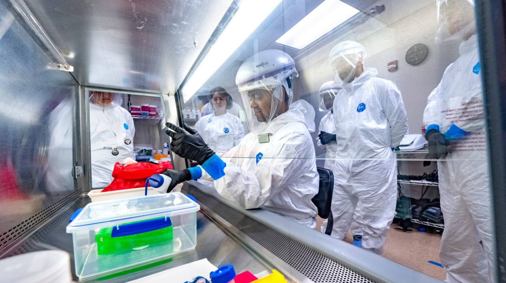 A group of students in PPE watch someone pour something behind protective glass