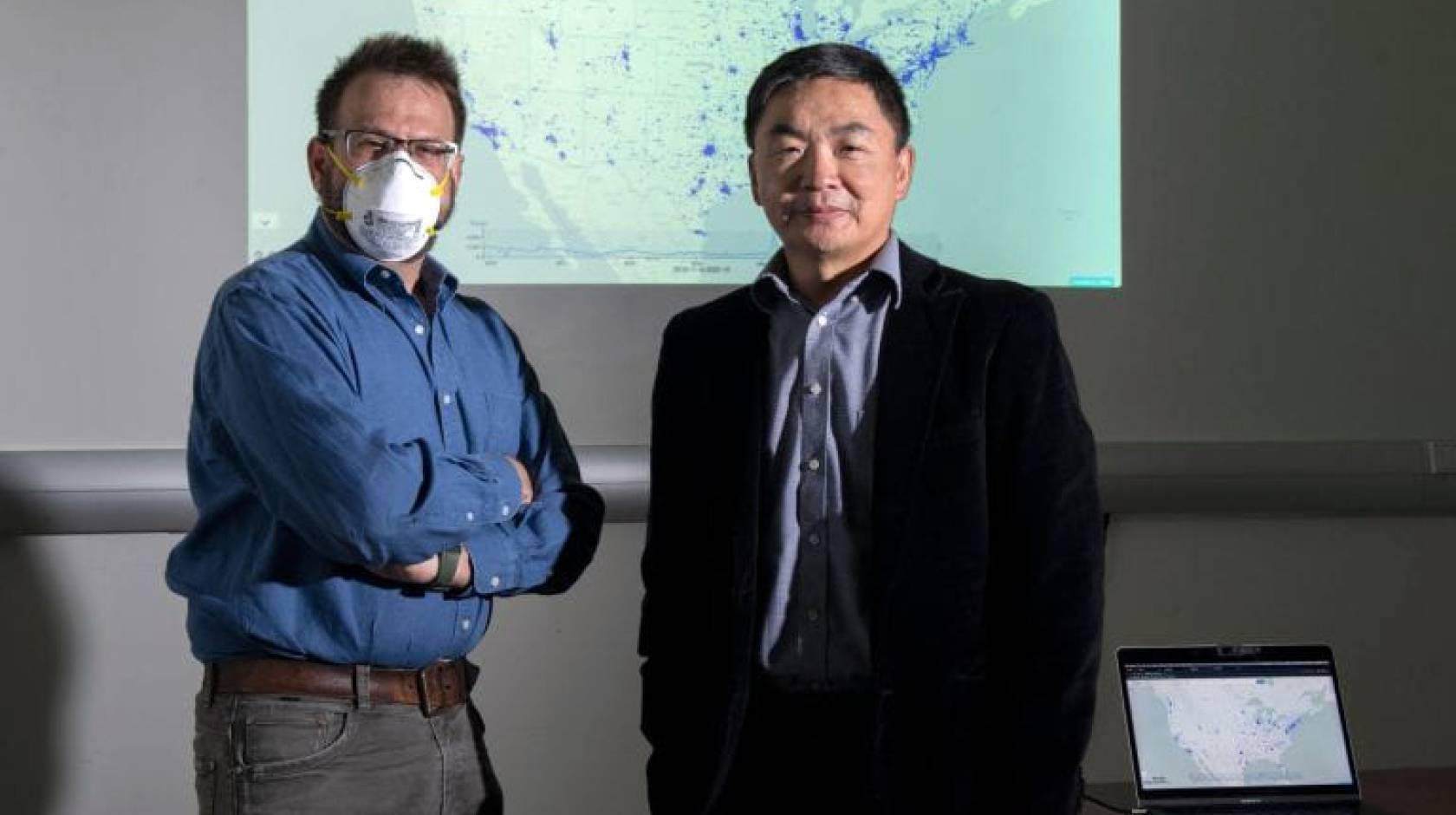 Two men standing together with a projector screen behind them, one wearing a face mask