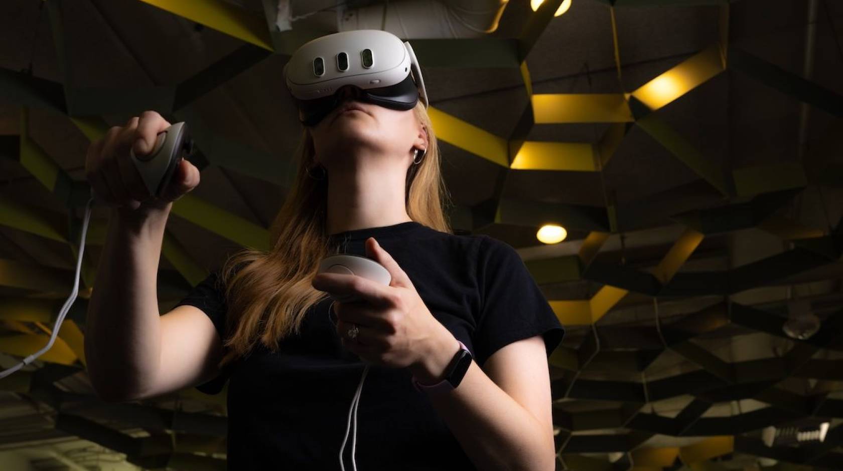 A student with a virtual reality headset and two controllers stands in a room with a honeycomb-esque roof