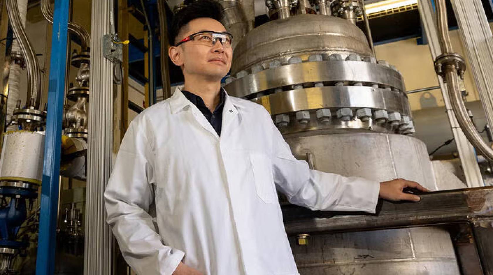 A professor in a white lab coat and safety goggles poses next to equipment